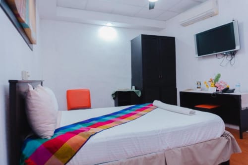 Habitación luminosa del Hotel Makarios en Tuxtla Gutiérrez con cama blanca, manta de rayas coloridas, TV de pantalla plana y diseño moderno