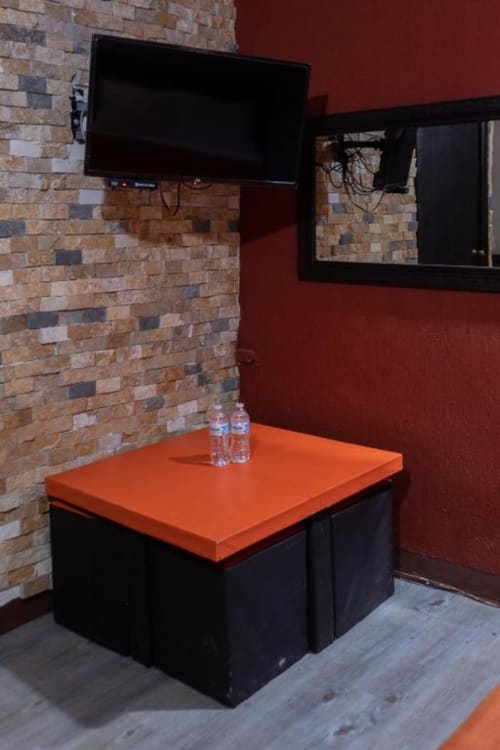 Rincón moderno de habitación en Hotel Makarios con mesa naranja, pared de ladrillo, TV montada y botellas de agua