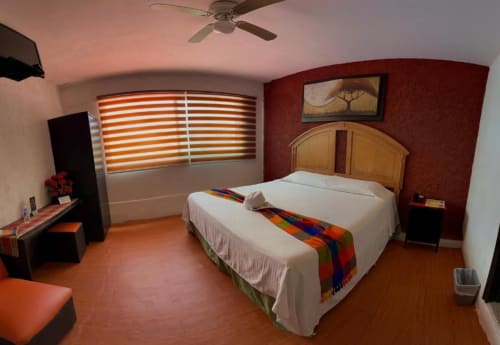 Habitación del Hotel Makarios en Tuxtla Gutiérrez con cama de madera, cortinas naranjas a rayas y pisos de terracota
