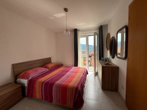 Camera da letto moderna nell'appartamento di Sormano con biancheria a righe rosse, balcone con vista sulla montagna, mobili in legno e illuminazione a sospensione.