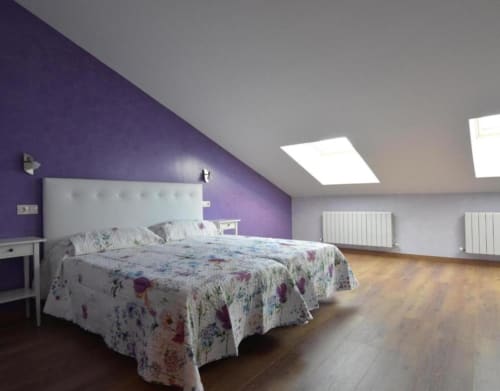 Moderno dormitorio abuhardillado en cabaña Nieva con pared de acento púrpura, cabecero blanco, ropa de cama floral, tragaluces y radiadores