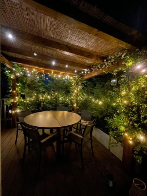 Elegante zona ristorazione su terrazza GIRESIO 41 Milano con tavolo rotondo, edera rampicante, luci sospese e pergolato in legno di notte