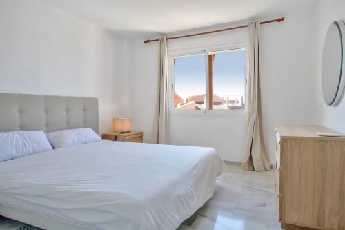 Dormitorio moderno y luminoso en el apartamento SACORO de Marbella con vistas al mar, ropa de cama blanca y cabecero beige