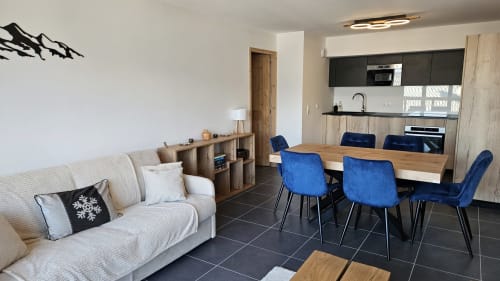 Séjour moderne à L'écrin des sapins avec canapé crème, chaises de salle à manger bleues, table en bois et cuisine équipée