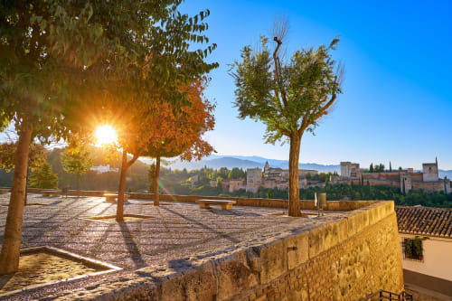 Mirador de San Nicolás en Granada con vistas a la Alhambra, árboles otoñales y el sol brillando al atardecer.