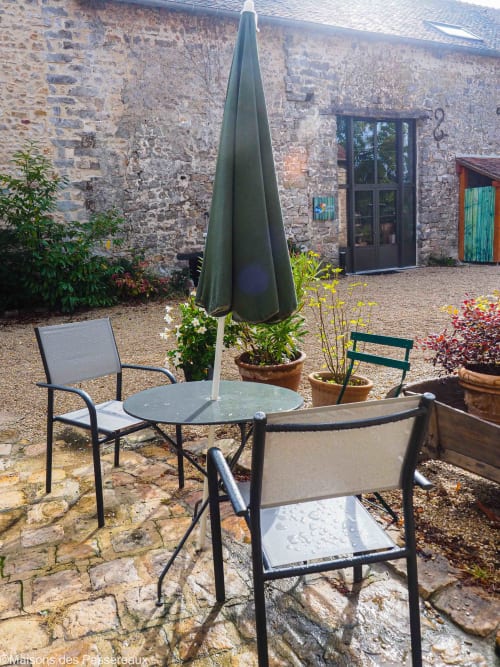 Terrasse ensoleillée gîte  Maisons des Passereaux : salon de jardin cosy sous parasol, murs en pierre authentiques, fleurs colorées, ambiance détente en forêt des Trois Pignons