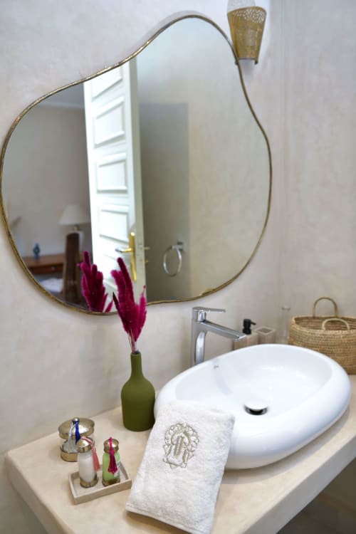 Elegante badkamer met golvende bronzen spiegel, witte wastafel, handdoek en roze bloemen in Riad Palau Marrakech.