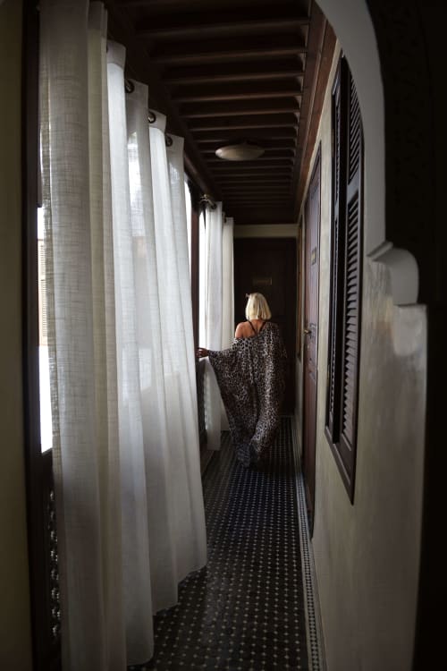 Vrouw in luipaardjas wandelend door een schaduwrijke gang met witte gordijnen in boutique hotel Riad Palau Marrakech