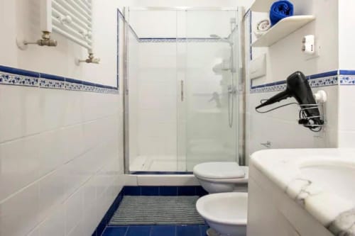 Bagno moderno con piastrelle bianche, dettagli blu, cabina doccia in vetro, doppio lavabo e toilette nell'appartamento di Roma