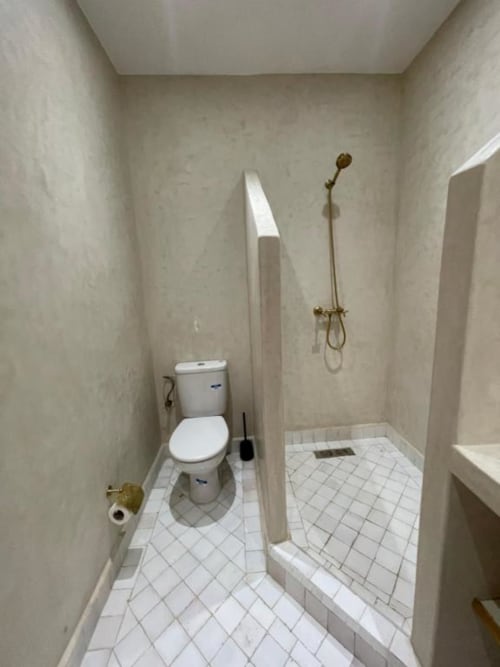 Salle de bains compacte du Riad Madinina avec carrelage blanc, toilettes et robinetterie de douche en bronze à Marrakech