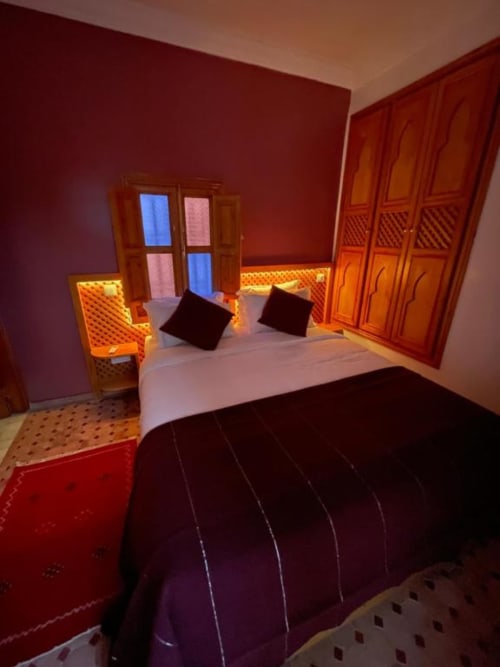 Chambre du Riad Madinina à Marrakech aux murs rouge chaleureux, portes en bois, tête de lit lumineuse et coussins foncés