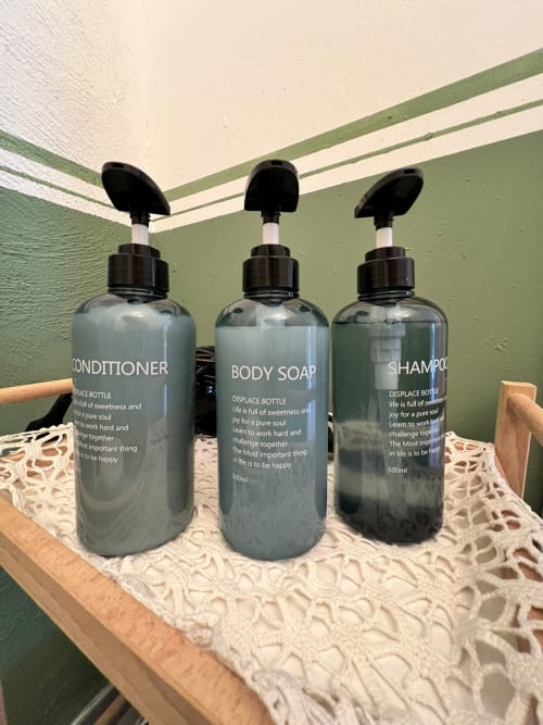 Three black pump dispenser bottles with conditioner, body soap, and shampoo labeled bottles on wooden shelf at La Cascina di