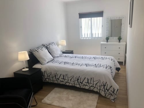 Quarto moderno em Sintra Apartamentos com cama de casal, criado-mudo preto, cômoda branca, espelho, luminária e assoalho de madeira