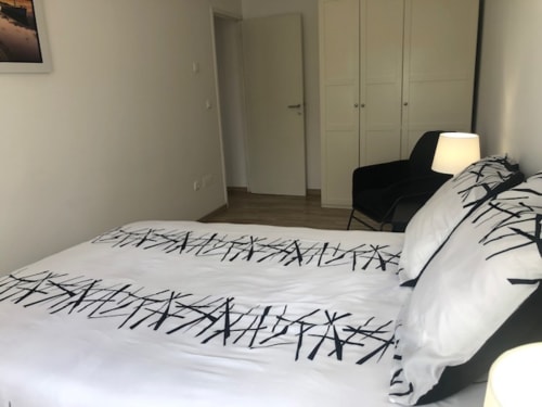 Quarto moderno no Sintra Apartamentos com roupa de cama branca com padrão de ramos pretos, cadeira preta, luminária e guarda-roupa