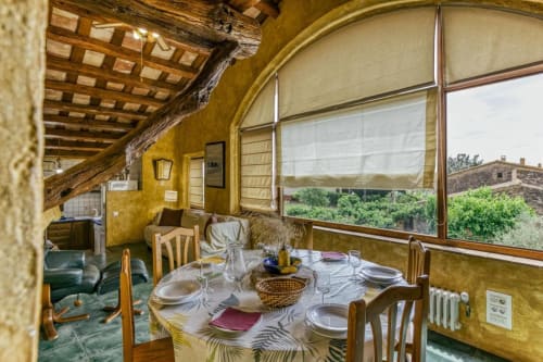 Comedor rústico en Can Gat Vell con vigas vistas, paredes amarillas, mesa redonda y ventanales panorámicos con vistas al Empordà