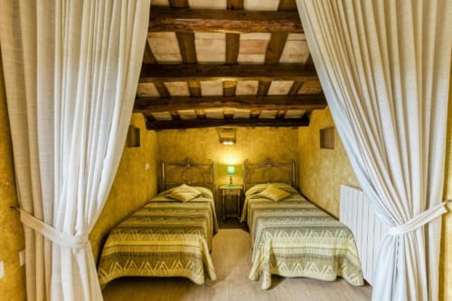 Dos camas gemelas en habitación catalana rústica con vigas vistas, paredes amarillas y cortinas blancas en Can Gat Vell, Girona