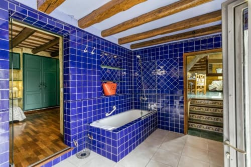 Baño de lujo con paredes de azulejos azul vibrante, vigas de madera, bañera, ducha de cristal y puertas verdes de madera en Can Gat Vell