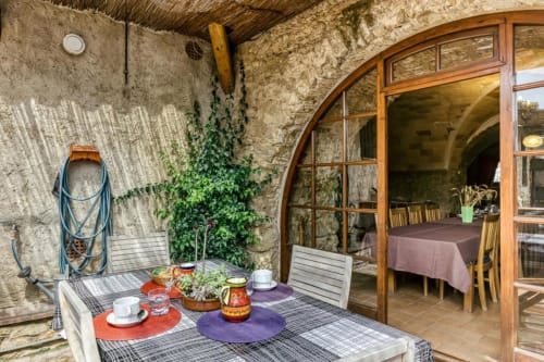 Terraza de Can Gat Vell en Girona con muros de piedra, mesa de comedor y puertas de madera arqueadas que conducen al comedor interior.