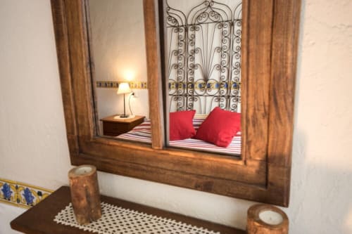 Espejo decorativo reflejando dormitorio con cabecera de hierro forjado ornamentado, almohadas rojas y lámpara en cabaña de Girona.