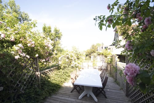 Table de salle à manger en plein air sur terrasse en bois à Vacances en Touraine, encadrée par des roses roses en fleur et une verdure luxuriante
