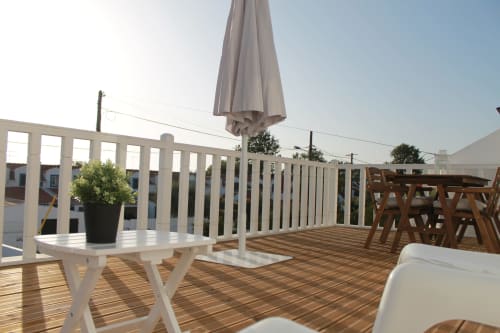 Varanda do apartamento Recantus Comporta com guarda-corpo branco, pavimento de madeira, guarda-sol, móveis de jantar e vista para o jardim