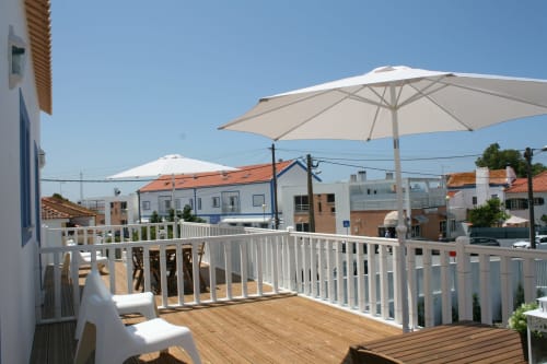 Varanda do apartamento Recantus Comporta com deck de madeira, guarda-corpo branco, guarda-sol e vistas da vila costeira