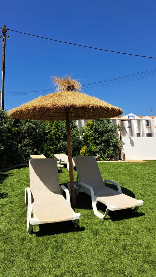 Dois espreguiçadeiras sob guarda-sol de colmo em jardim exuberante no Recantus Comporta, Comporta, Portugal