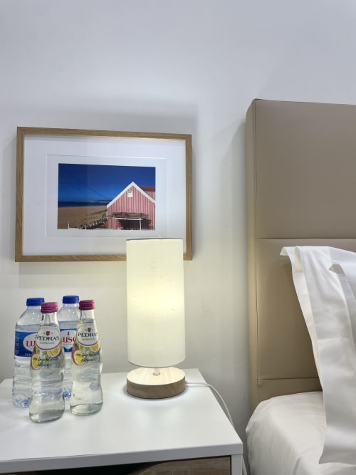 Vila Alexandra hostel room with framed coastal barn photo, bedside lamp, water bottles, and white linens in Costa da Caparica