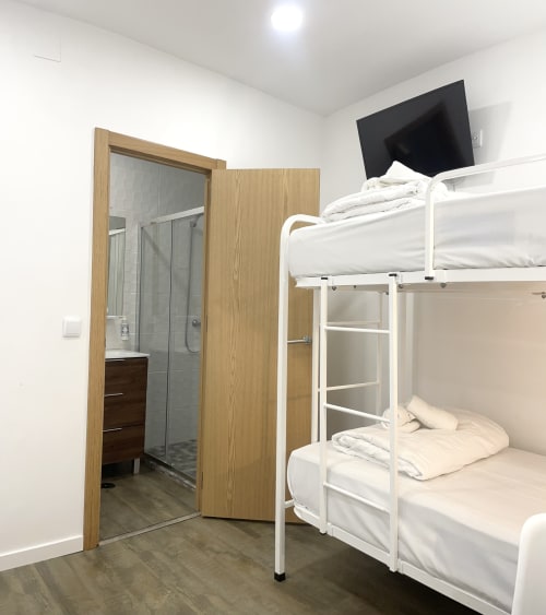 Modern hostel room at Vila Alexandra, Costa da Caparica with white bunk beds, TV, wooden bathroom door, and bright white