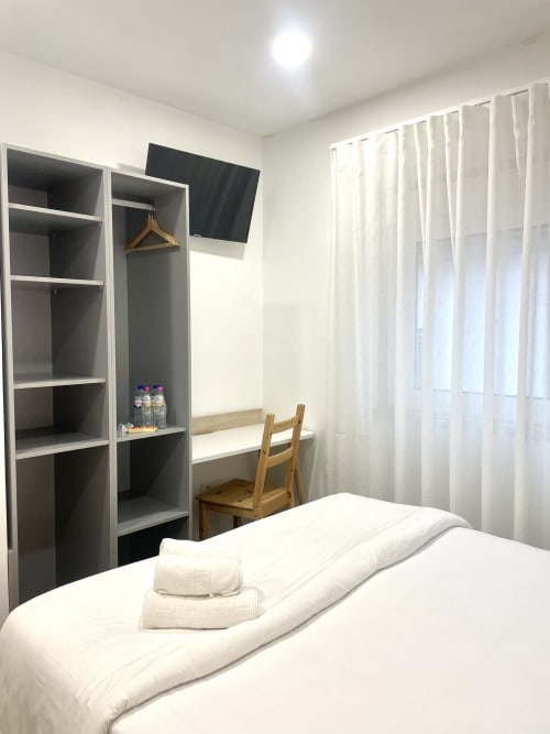Modern hostel bedroom at Vila Alexandra Costa da Caparica with bed, shelving, desk, chair, TV and sheer curtains