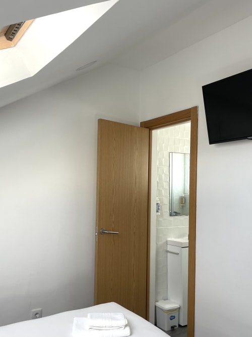 Modern hostel room at Vila Alexandra in Costa da Caparica with wooden door, bathroom entrance, TV, and white bed with towels