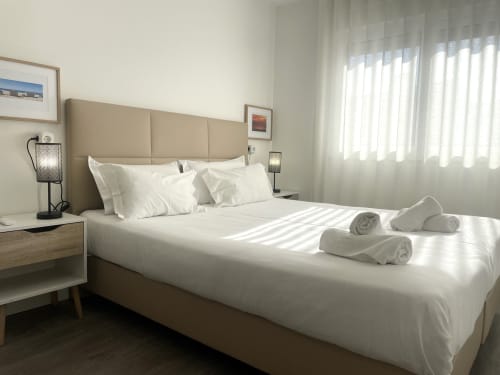 Modern bedroom at Vila Alexandra hostel in Costa da Caparica with beige headboard, white linens, rolled towels, and natural