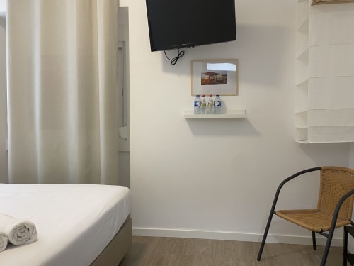 Modern hostel room at Vila Alexandra in Costa da Caparica with bed, TV, white shelving, and wicker chair