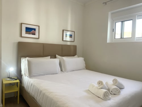 Bright bedroom at Vila Alexandra hostel in Costa da Caparica with rolled towels on bed, taupe headboard, yellow nightstand,