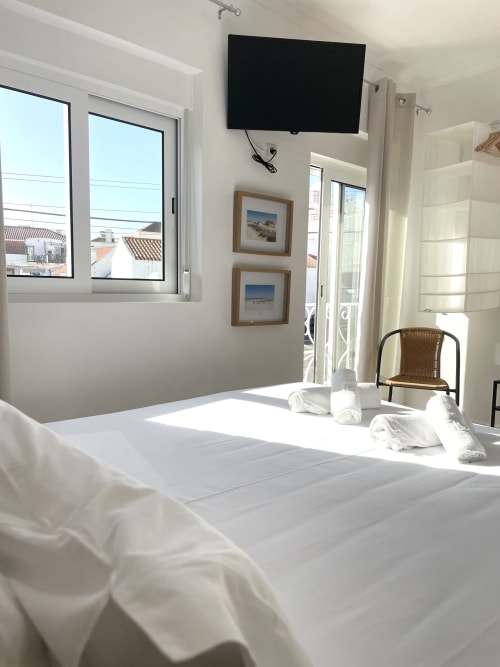 Bright hotel room at Vila Alexandra in Costa da Caparica with white bed, wall-mounted TV, framed beach art, and city view