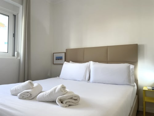 Bright modern hostel room at Vila Alexandra in Costa da Caparica with white bedding, rolled towels, tan headboard, and city