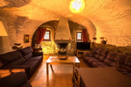 Sala de estar rústica de piedra en Girona con chimenea, techos abovedados, suelos de barro y muebles a cuadros.