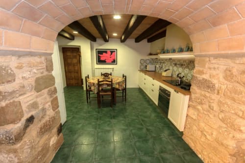 Cocina rústica de piedra con suelo de azulejos verdes, vigas de madera, mesa de comedor y armarios color crema en Can Gat Vell
