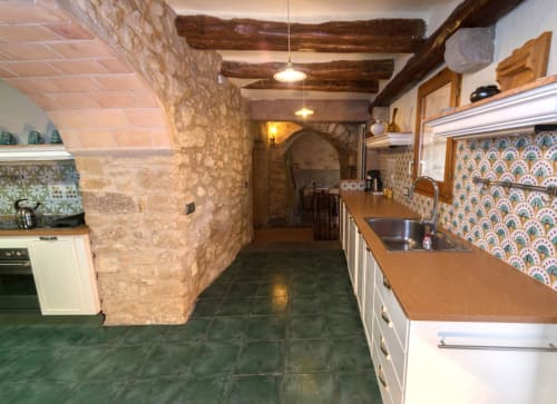 Cocina rústica de piedra con baldosas terracota, vigas de madera, azulejos patterned y vistas al jardín de Can Gat Vell,