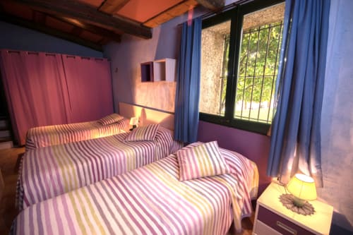 Dormitorio luminoso con ropa de cama rayada en la casa Can Gat Vell en Girona, ventana con rejas y vista al jardín, atmósfera cálida