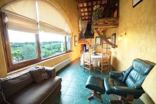 Salón luminoso con sillones de cuero negro, comedor y gran ventanal arqueado con vistas a la campiña de Girona en Can Gat