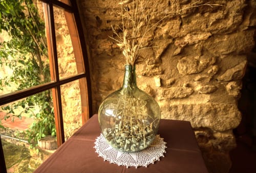 Flores secas en botella de vidrio sobre paño de encaje junto a la ventana de la casa Can Gat Vell, Girona, con pared de piedra de fondo