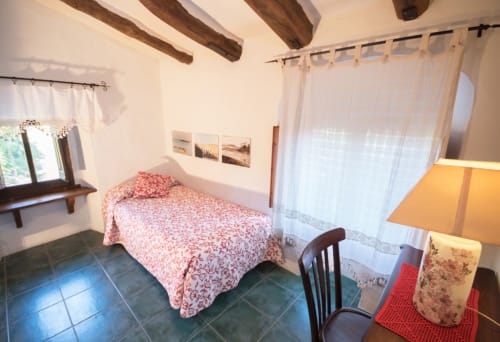 Dormitorio luminoso con colcha floral roja, vigas de madera, baldosas de terracota y cortinas blancas en la casa rural Can Gat Vell, Girona