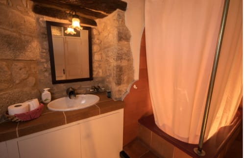 Baño rústico de la casa rural Can Gat Vell en Girona con muros de piedra, lavabo blanco, espejo y cortina de ducha