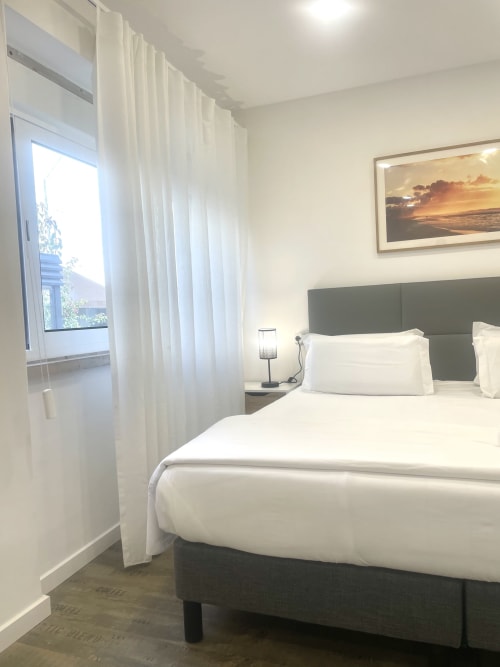 Modern bedroom at Vila Alexandra hostel in Costa da Caparica with white bedding, gray headboard, and city view through sheer