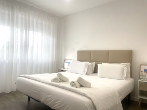 Bright, modern bedroom at Vila Alexandra hostel in Costa da Caparica with white bedding, beige headboard, and sheer curtains