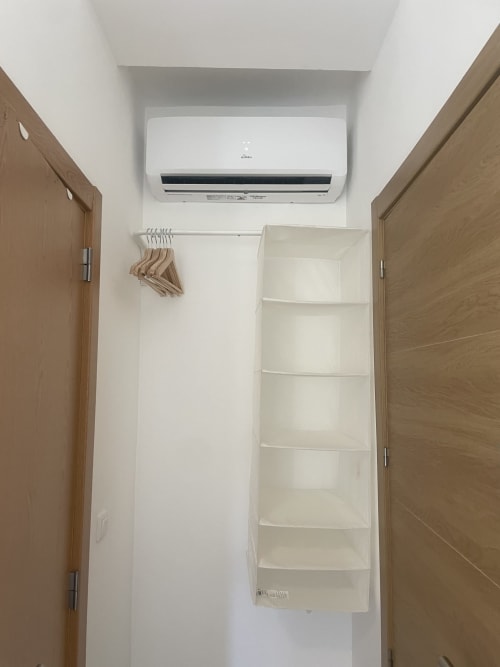 Narrow hostel room with white walls, wall-mounted air conditioning unit, wooden doors, and built-in shelving at Vila