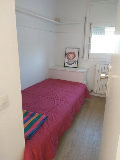 Dormitorio compacto en Finques Garcia Enrich, Palafrugell con cama burdeos, cabecero rosa, paredes blancas, radiador y ventana.