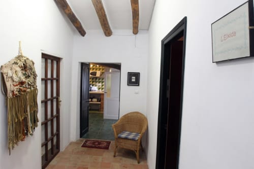 Pasillo rústico en Can Gat Vell B&B en Girona con vigas vistas, silla de mimbre y macramé decorativo en la pared.