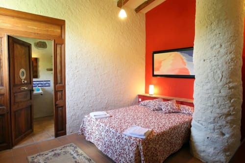 Acogedor dormitorio en la casa rural Can Gat Vell de Girona con pared roja, ropa de cama estampada y detalles de piedra rústica.